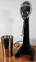 Side view of milkshake maker and stainless steel cup showing chrome head and black base.