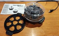 Full view of Hamilton Beach ezEgg cooker with clear dome lid and 7-slot egg holder tray beside it, along with instruction manual.