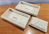 Two rectangular light-colored marble trays and one marble soap dish shown on wooden surface with visible natural veining in marble.