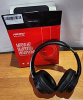 Black MPOW H7 Bluetooth headphones with cushioned ear pads in front of original red and black box featuring the product name and logo, placed on wooden surface.
