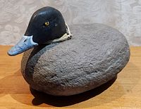 Side angled front view of the stone folk art duck showing the gray stone body, black painted wooden head, and blue-gray beak with painted yellow eye.