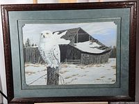Framed art print showing a snowy white owl perched on a fence post with a rustic barn covered with snow in the background.