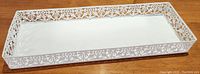 White rectangular metal decorative tray showing intricate cutout pattern on the raised edges, placed on a wooden surface.