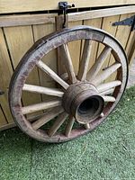 Wooden wagon wheel leaning against wooden fence on artificial grass with visible spokes and metal band around circumference.