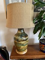 Full lamp with burlap lampshade resting on a wooden table, showing ceramic base with earthy swirled colours of green, chocolate, sand, and cinnamon.