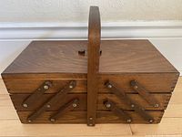 Closed view of the wooden sewing box showing the handle and the side hinged tiers with metal pivot arms.