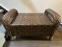 Full view of rectangular wooden ottoman with woven wood surface and raised woven wooden armrests on both ends. Four woven legs support the structure.