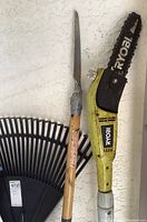 Close-up of a Ryobi electric tree trimmer with a black protective blade cover, a large black plastic rake, and a hand saw with a wooden handle wrapped in duct tape.
