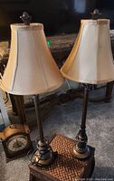 Two matching bedroom lamps side-by-side with beige fabric bell-shaped lampshades and decorative dark metal bases with finials on top of shades. Coiled power cords visible.