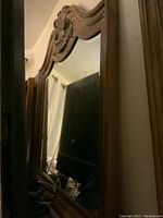 Photo of ornate wooden framed mirror with visible carved scrollwork at the top of mirror frame. Mirror glass reflects part of the room.