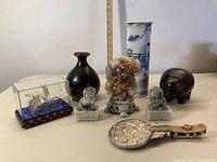 Frontal group shot showing black ceramic vase, marble Foo dogs, carved stone sculpture, porcelain vase, wooden pig, hand mirror and silver rickshaw figurine.
