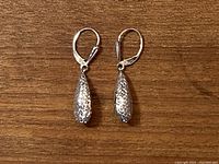 Pair of textured teardrop-shaped white gold dangle earrings with lever-back clasps on a wooden surface.
