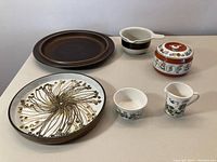 Photo showing overall ceramic lot with plates, bowl, pitcher, and lidded container arranged on table.