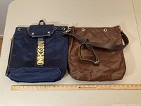 Photo of both bags side by side next to a ruler showing scale, the backpack in navy blue canvas and the brown leather crossbody bag.