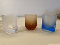 Three short frosted Moser Tipsy tumblers in clear, amber and blue, side by side
