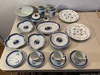 Overview of entire porcelain dinnerware lot showing blue and white floral patterned pieces including teacups, saucers, plates, ruffled dishes, creamers, sugar pot, platter, and large bowl arranged on table.
