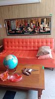 Orange vinyl couch with tufting and two pillows on it, placed in a room with a wooden framed painting above and a wooden coffee table in front with decorative items.
