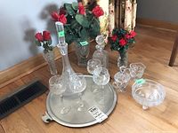 Wide angle photo showing the entire set including vases with artificial red roses, decanters, stemware, bowl, cream and sugar set, all displayed on a round metal tray on wooden floor.
