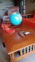 Photo showing a wooden table with two orange-red tone glass bowls, one orange-red glass ashtray, metal wall hanging, and Imperial blue-pink world globe on metal stand.