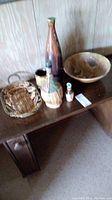 Photo of lot items including decorative bowl, basket, ceramic mug, and decorative bottles on a table against a wood-panel wall.