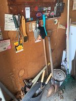 Wood-handled garden tools including shovels, rakes, and hand tools resting against a wall inside a shed.