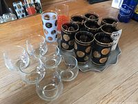 Overview of the glassware set showing 6 black bubble glasses with gold designs, one white transfer ware glass, one orange transfer ware glass, and the metal tray underneath.