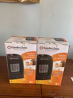 Front view of two boxed Comfort Zone ceramic oscillating portable space heaters showing branding, product images, and family photo.