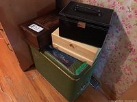 Wood and faux leather jewelry boxes stacked on green vinyl hamper against floral wallpaper and wooden floor
