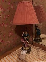 Vintage table lamp featuring ceramic female figurine base in blue and pink dress with fabric shade, photographed against floral wallpaper.