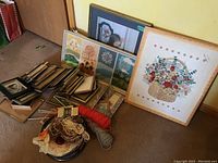 Photo showing framed needlepoint artworks, yarn skeins, crafting tools, books, and a pile of various empty wooden and black picture frames.