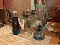 Two glass oil lamps shown on a striped cloth surface, one lamp with a blue base and tall hurricane shade, the other with a clear etched base and bronze tone burner, without shade.