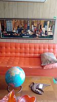 Front view of framed abstract city skyline print above orange couch with world globe and decorative objects on table below.