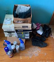 Overview photo showing stacked shoe boxes, various pairs of shoes including runners and boots, and multiple shoe care products on the floor.