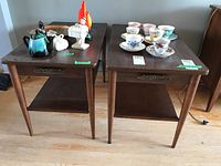 Pair of Mid-Century Modern side tables shown with decorative items on top. Wood veneer bases visible with dark laminate tops, faux drawer details with metal pulls, tapered legs, and lower shelf.