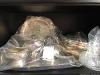 Multiple silver plated pieces of tea set and serving items wrapped in plastic bags, located on stainless steel tray, seen from front
