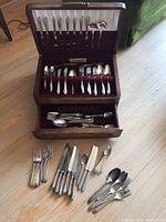 Full view of open wood flatware canteen showing two drawers stocked with silver plate and stainless steel cutlery and various loose pieces arranged in front.