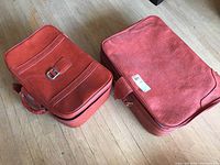 Two vintage red vinyl suitcases placed side by side on a wooden floor, showing their top surfaces and front buckle strap on smaller case.