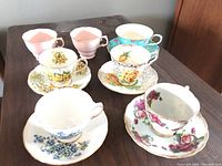 Overview of 7 teacups and saucers set with floral designs and two plain pink orphan cups