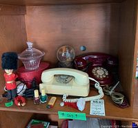 Full shelf view showing white and burgundy rotary dial telephones, musical snow globe, glass candy dish, British soldier figurine, nail polish, lighter, keychain, and other small items.