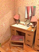 MCM wood vanity with attached large rectangular mirror and four drawers with metal pulls. Vanity surface has a cloth runner, two pink lamps, and decorative items. Matching wooden stool with red cushioned seat in front of vanity.