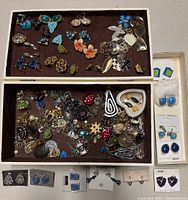 Two trays filled with a large variety of vintage earrings with different styles (screwback, clip, post), colors, and designs, displayed along with some packaged earrings.