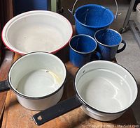 Top-down view of all enamelware including blue mugs, blue pot, white bowl, and two white sauce pots on wooden surface.