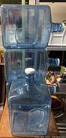 Four stackable 3-gallon plastic water bottles with white lids and handles, translucent blue color, embossed "3 GALLONS" marking.