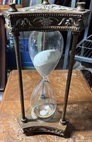 Front view of the hourglass showing glass bulbs with white sand and the antique brass-colored ornate metal frame.