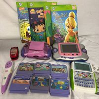 Photo of LeapFrog handheld consoles, Tag Reader pen, learning activity books, and game cartridges showing all items included in the lot