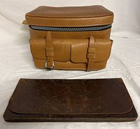 Tan Belding leather satchel with two front buckle straps and zippered top, sitting behind a dark brown vintage leather wallet with surface wear
