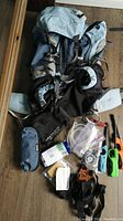 Top view of survival backpack laid out with survival accessories spread around it including hydration system, lighters, knife, compass, whistle, fire starter, paracord, and pouches.