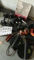 Photo showing mixed fitness equipment including 15 and 10 lb dumbbells, Everlast pushup handles, skipping rope, weight straps, braces, and exercise guides.