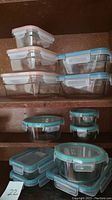 Twelve assorted glass food storage containers stacked in a cabinet, showing various rectangular and round shapes with light blue and light pink snap lids.