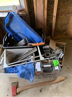Top view of blue Rubbermaid bins filled with various hardware and tools including a hand saw, Teflon tape, screws, and an axe placed on the floor.
